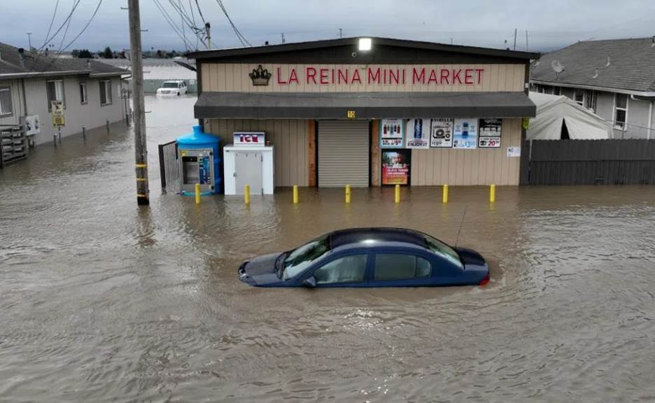 A black car sits in front of a small building with a sign reading ‘La Reina mini market’, both are submerged in flood waters.