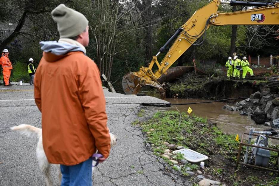 A man watches as workers operate an excavator on a washed-out road.