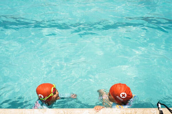 A view from a pool’s edge looking down on two children holding onto the pool wall. Both children are wearing red-orange swimming caps that bear the American Red Cross logo.