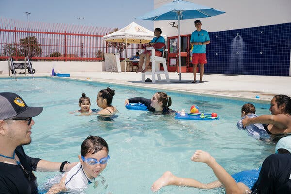 Parents, children and swim instructors swim in the pool while two lifeguards, one seated and one standing, in blue shirts and red shorts, watch over.