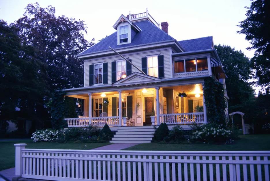 A home with lights on on the porch and a white picket fence surrounding the property