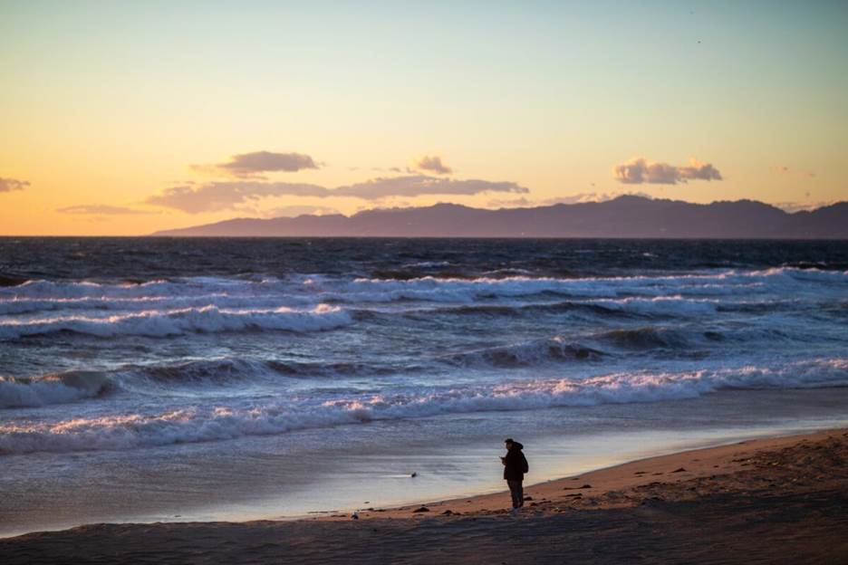 A lone small figure stands on the shore of a beach at sunset.