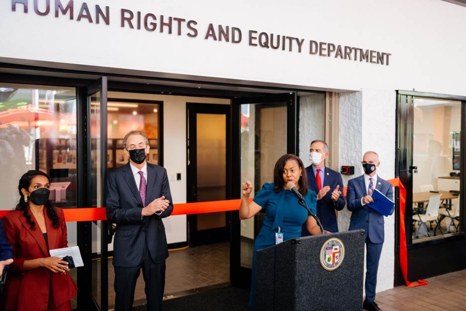 A woman is standing at a podium in front of a building with the words Human Rights and Equity Department on it.