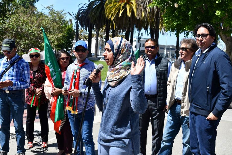 A person speaking into a microphone with a group of people standing in the background  Description automatically generated with low confidence