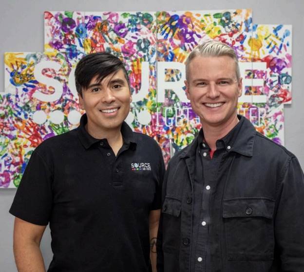 Two men standing in front of a colorful wall  Description automatically generated