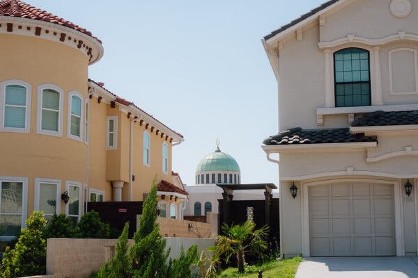 Two houses sit side by side, one that is yellow-ish on the left, and one that is light-colored that features two garage doors. In the distance, behind the homes, is a domed building.