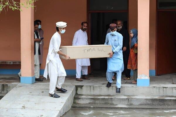 Two men in surgical masks carry a plain coffin out of a building as several other people look on.