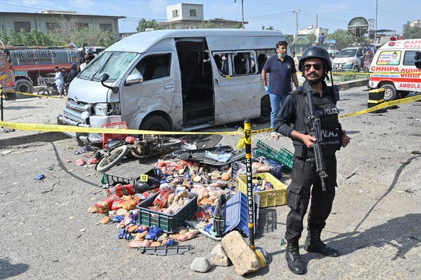 A police officer in riot gear, holding an assault rifle, stands in front of a roped-off crime scene that includes a damaged van, a motorcycle on its side and a spilled load of groceries.