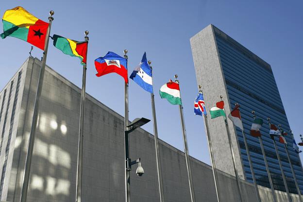 A group of flags on poles with Headquarters of the United Nations in the background Description automatically generated