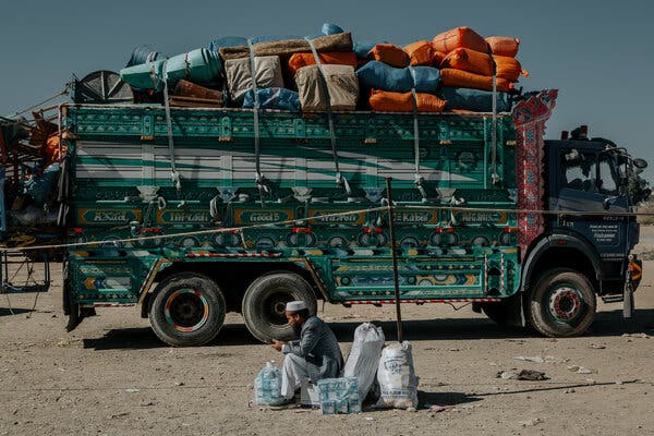 A person sits on dusty ground next to bottles of water and white sacks. A large green truck, brightly painted and heavily loaded with colorful bundles, is behind them.