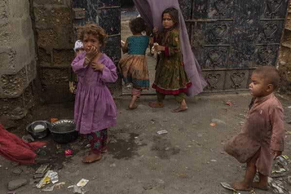 Four barefoot children stand on dusty ground in front of a dark, ornate metal gate. A child in a light-brown tunic stands in the foreground, facing away.